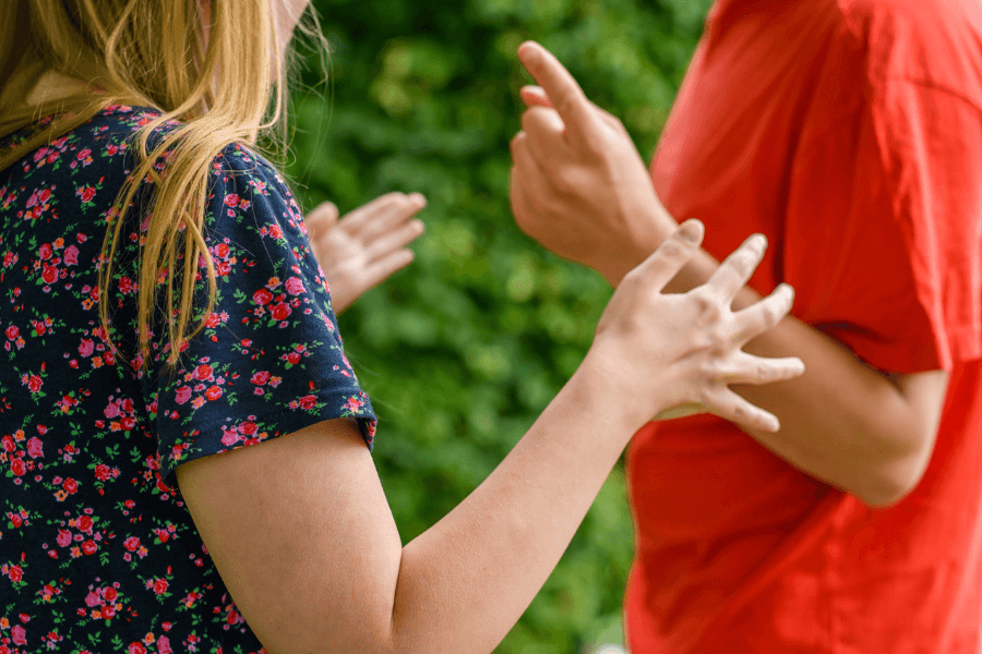Close-up of two people in a heated argument, with expressive hand gestures and tense body language, standing outdoors against a backdrop of green foliage.