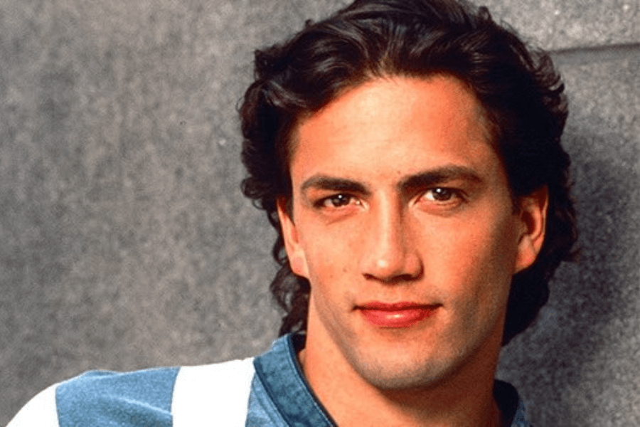 This image features a man with medium-length dark hair and a slight smile, wearing a casual blue shirt with white accents. The background is a neutral gray surface, emphasizing a clean and simple portrait-style composition.