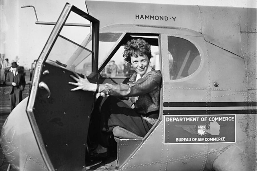 A black-and-white photograph of a woman sitting in the cockpit of a small aircraft, smiling as she reaches for the door. The plane has a sign that reads "Department of Commerce, Bureau of Air Commerce," and the model name "Hammond-Y" is visible above the window. In the background, a man in a suit and hat stands observing the scene.