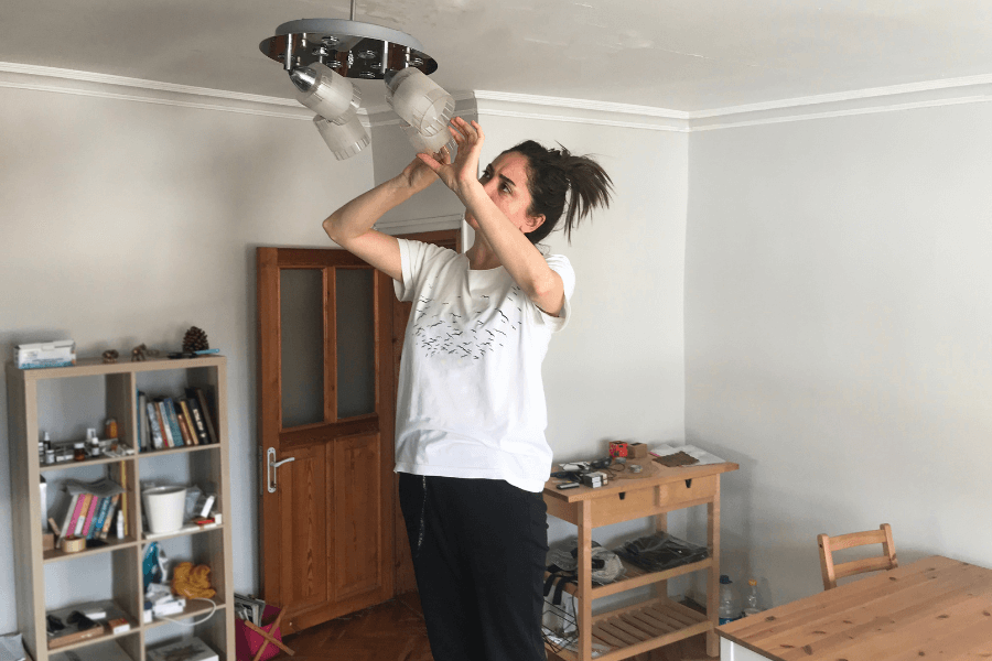 This image shows a woman standing on a surface in a room, reaching up to adjust or fix a ceiling light fixture. She is wearing a casual white T-shirt and dark pants, with her hair tied back. The room features minimal furniture, including a bookshelf, wooden door, and table, emphasizing a home environment.