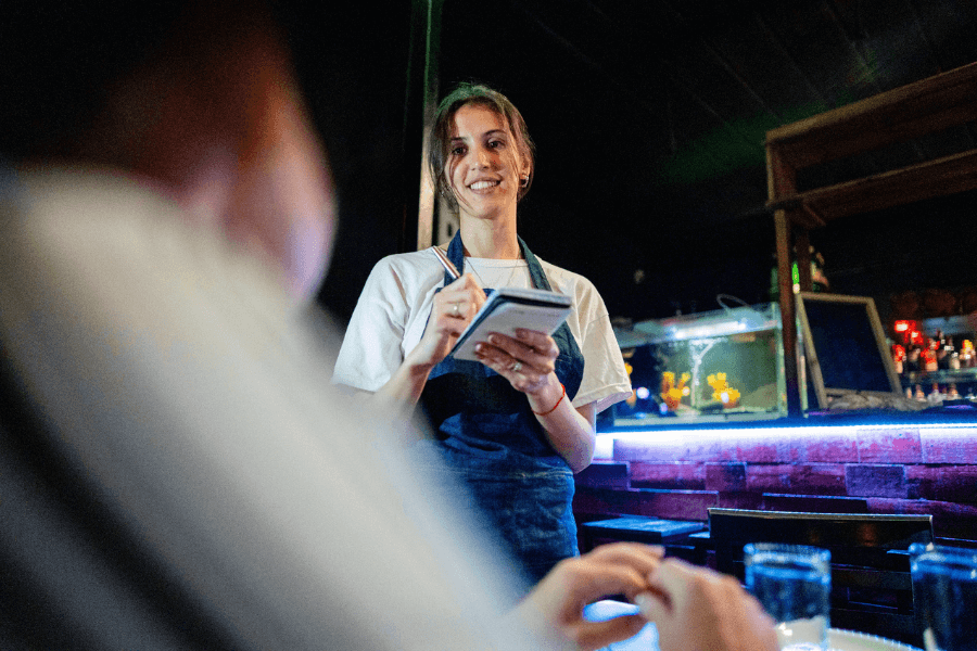Smiling waitress in a white t-shirt and apron taking an order with a notepad inside a dimly lit restaurant, interacting warmly with a customer.