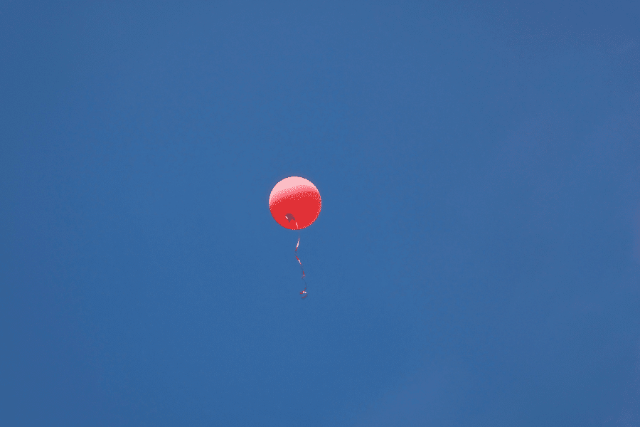 This image shows a single red balloon floating against a clear blue sky. The balloon's ribbon dangles freely, adding a whimsical and carefree touch to the composition.