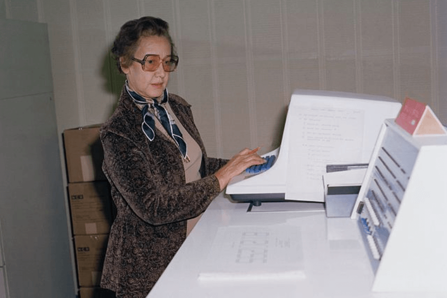 A woman wearing glasses and a patterned blazer stands at a white desk, working on an early computer. She types on a blue keyboard while reviewing a printed document, with a box of punch cards and additional equipment nearby. The setting appears to be an office or laboratory, with stacks of cardboard boxes in the background.