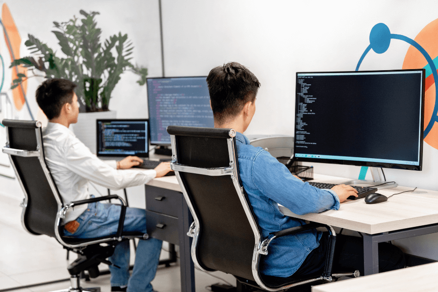 This image shows two individuals seated at desks, working on computers in an office environment. Both are viewing large monitors displaying lines of code, suggesting a programming or software development activity. The workspace includes modern office chairs, a plant in the background, and colorful wall decor, giving a tech-savvy and collaborative vibe.