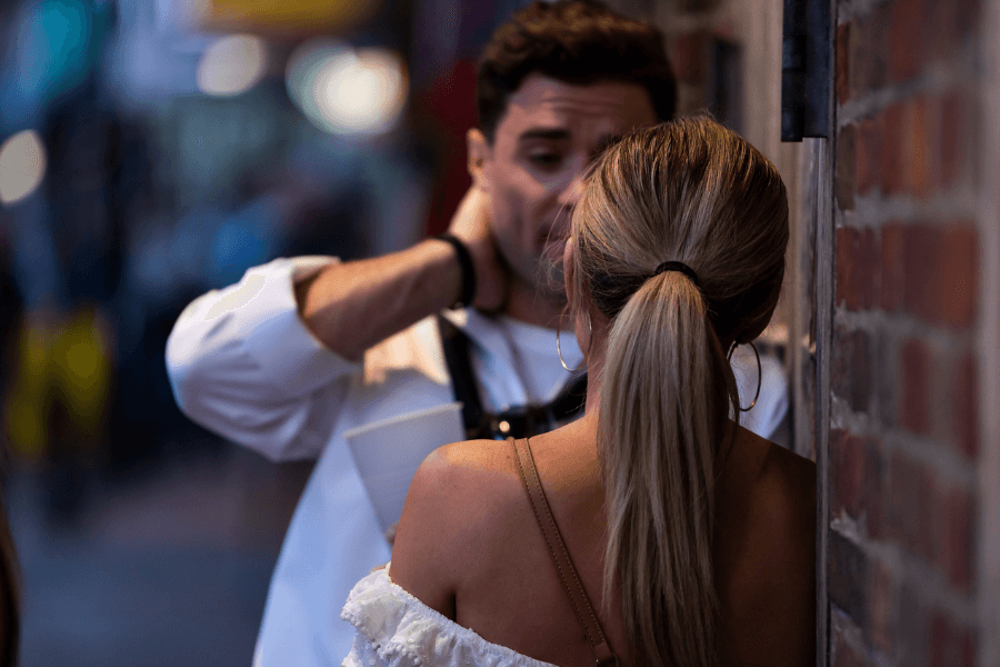 Man and woman engaged in a serious or emotional conversation in a narrow alley or street at dusk, with the woman facing away from the camera and the man appearing distressed or thoughtful.