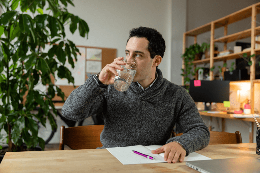 A man in a gray sweater sits at a wooden desk in a modern office, drinking a glass of water. Papers, a pen, and a laptop are on the desk, while shelves with plants and office supplies are visible in the background.