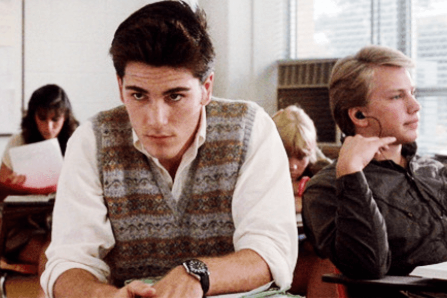 This image shows a young man sitting in a classroom, wearing a patterned sweater vest over a white shirt and looking focused or pensive. Another student, seated beside him, appears more relaxed, wearing headphones and resting their hand on their face. The background includes other students engaged in classroom activities, along with windows providing natural light.