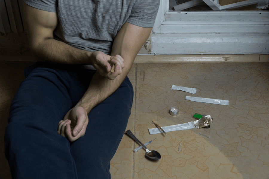 Man sitting on the floor preparing to inject drugs, surrounded by paraphernalia including syringes, a spoon, and foil, highlighting a scene of substance abuse and addiction.
