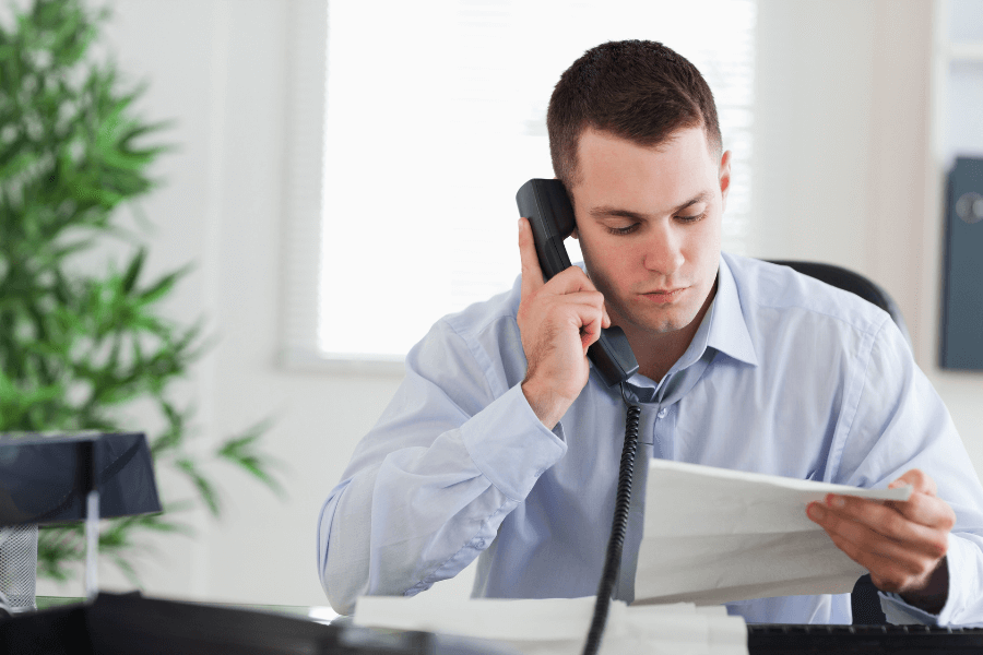 This image shows a man in a professional setting, sitting at a desk while speaking on a landline phone. He is holding and reviewing a sheet of paper, appearing focused and engaged in the task. The office environment includes natural light and a plant in the background, creating a productive atmosphere.