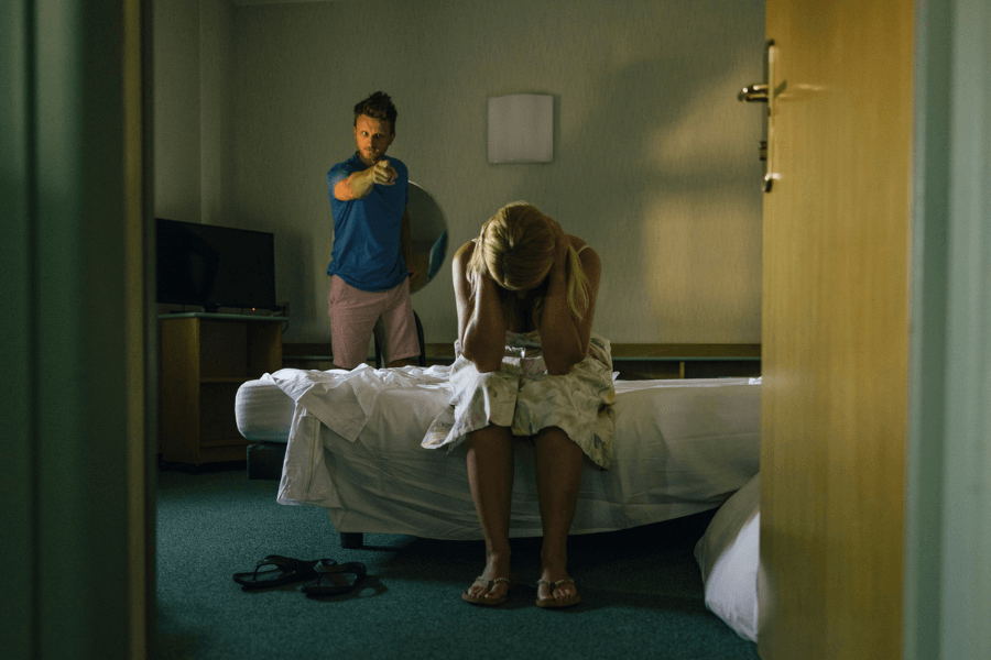 Man standing and pointing aggressively at a woman who is sitting on a bed with her head in her hands, appearing distressed, in a dimly lit room—depicting a scene of emotional or verbal abuse.