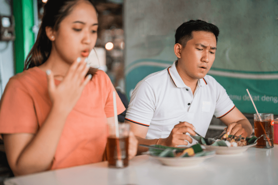 A woman in an orange shirt fans her mouth while holding a drink, seemingly reacting to spicy food, as a man in a white polo shirt looks at his plate with a concerned expression. They are dining at a casual restaurant with traditional food served on banana leaves.
