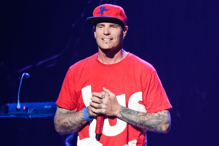 This image shows a man wearing a red t-shirt and a matching red cap with a blue graphic design. He has tattoos on his arms and is holding a microphone while standing on stage. The background is dimly lit with a performance setting, creating a vibrant and energetic atmosphere.
