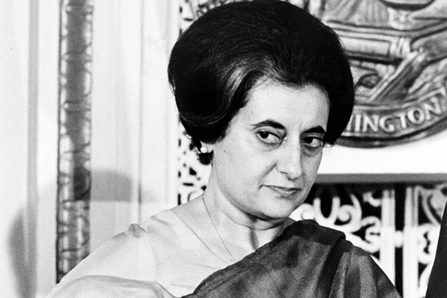 A black-and-white photograph of a woman wearing a sari, with a serious and contemplative expression. Her distinctive hairstyle is neatly styled, and she appears to be in a formal setting with an official seal visible in the background. The lighting and composition emphasize her strong gaze and presence.