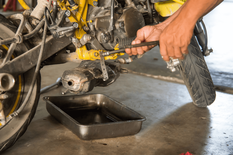 This image shows a person working on a motorcycle, using a tool to adjust or repair a component near the engine. A black pan is placed beneath the bike, likely to catch oil or other fluids during maintenance. The setting appears to be a garage or workshop, with visible motorcycle details emphasizing hands-on repair work.