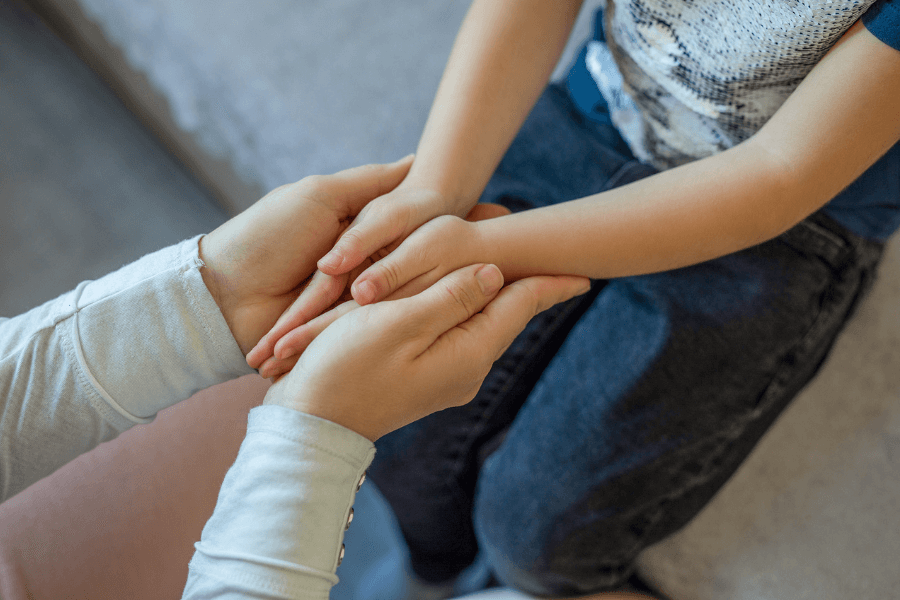 Close-up of an adult gently holding a child's hands, capturing a moment of comfort, support, or reassurance, with the child seated and partially visible.