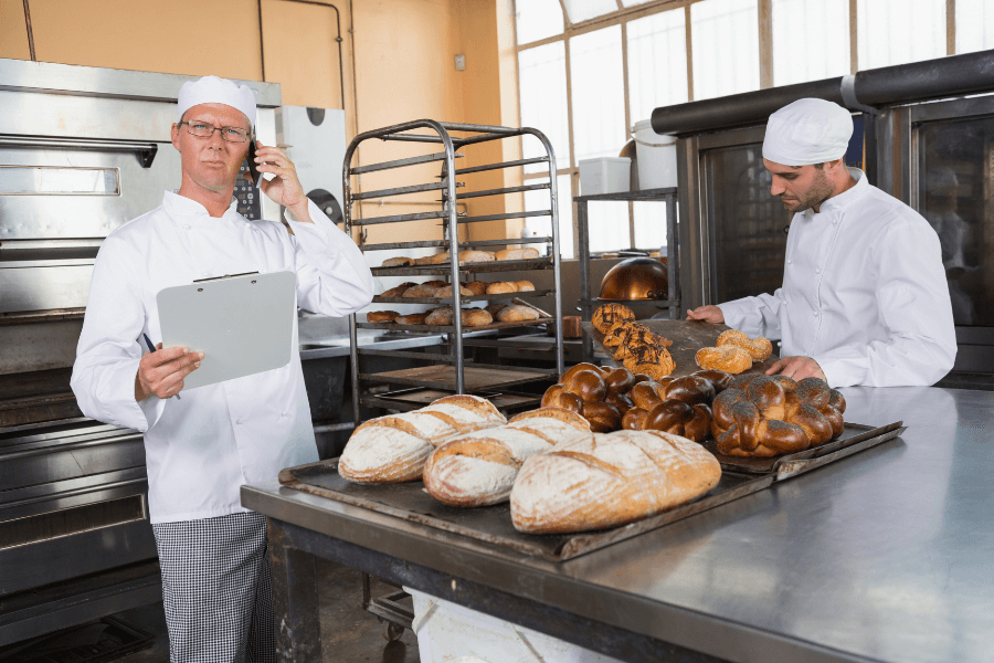 This image shows a professional bakery setting where two bakers are at work. One baker is talking on the phone while holding a clipboard, and the other is arranging freshly baked bread and pastries on a tray. The countertop is filled with an assortment of artisan loaves, and racks of baked goods are visible in the background.