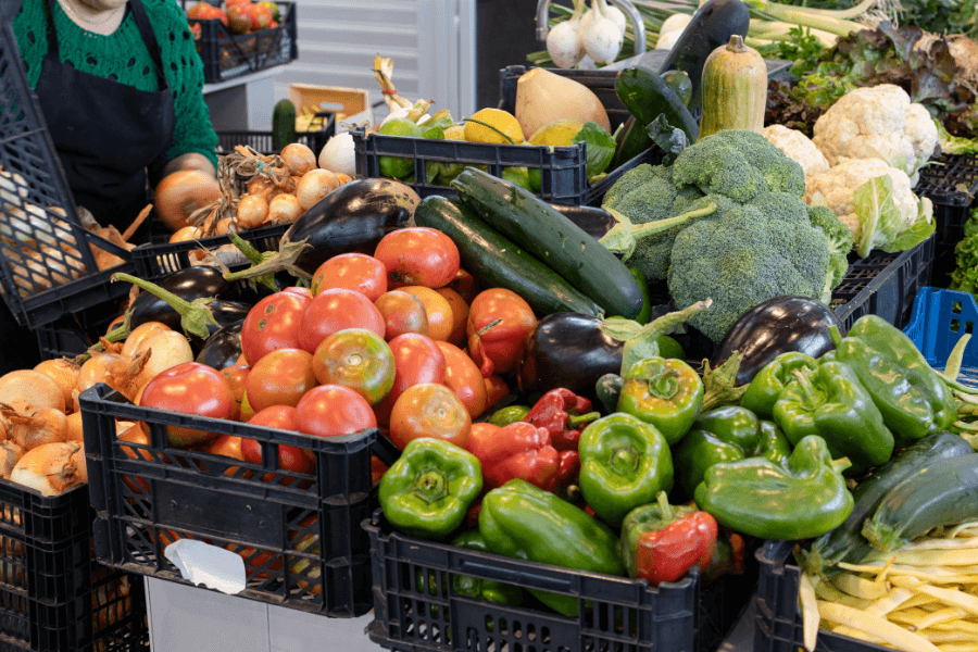 A colorful display of fresh vegetables at a market stand, including tomatoes, bell peppers, cucumbers, eggplants, onions, and broccoli. A vendor in a green sweater and black apron is seen handling produce in the background.