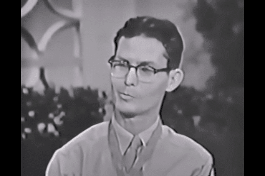 Black-and-white still from a television interview showing Desmond Doss wearing glasses and a collared shirt, with the Medal of Honor around his neck. He appears calm and composed while speaking, with a softly lit background featuring plants and abstract patterns.