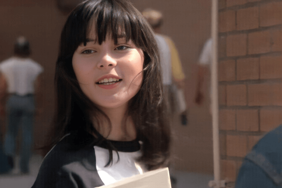 This image features a young woman with dark hair and bangs, smiling as she looks off to the side. She is wearing a black and white shirt and holding what appears to be a notebook or folder. The setting is outdoors near a brick wall, with a soft background of other people in casual clothing, suggesting a school or campus environment.