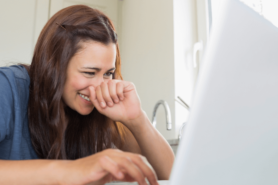 This image shows a woman sitting in front of a laptop, smiling and appearing amused or entertained by something on the screen. She has her hand partially covering her mouth, adding a playful or bashful expression. The background includes a bright and airy kitchen setting, enhancing the casual and relaxed atmosphere.