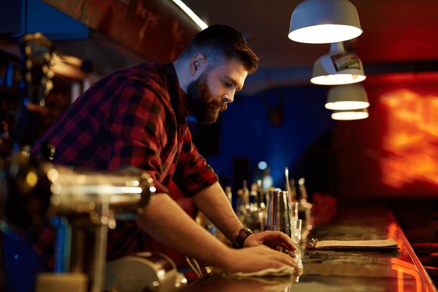 A bartender in a red plaid shirt leans over the bar, wiping the counter with a cloth in a dimly lit bar. The setting features hanging lights, drink shakers, and taps, with a warm, vibrant background glow.