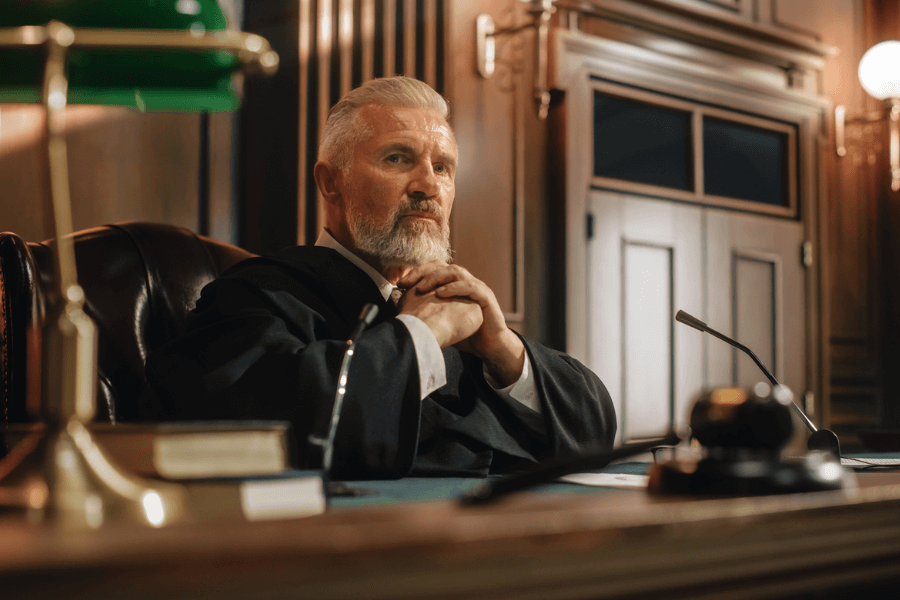 Serious-looking male judge with white hair and beard seated in a courtroom, hands clasped under his chin, surrounded by legal documents, a gavel, and a desk lamp.