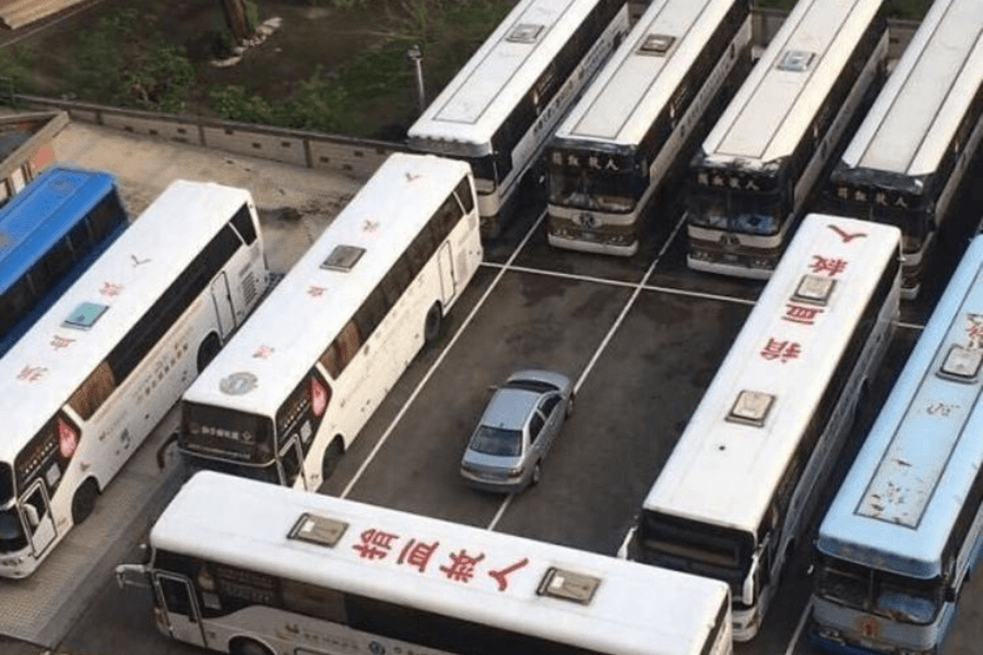 This image shows a small silver car parked in a lot surrounded and boxed in by several large buses, making it impossible for the car to leave. The buses are arranged closely in a seemingly deliberate manner, creating a striking and humorous visual of the car trapped in place.
