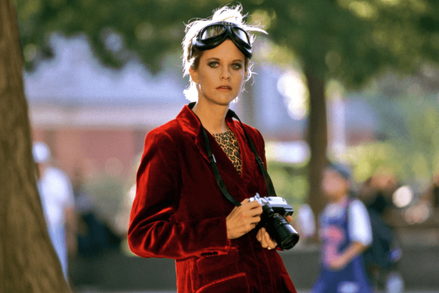 This image depicts a woman wearing a vibrant red velvet jacket, leopard-print top, and goggles resting on her head. She is holding a camera, suggesting she is a photographer or observer, with a composed and confident expression. The background is an outdoor setting with blurred trees and people, creating a dynamic and stylish atmosphere.