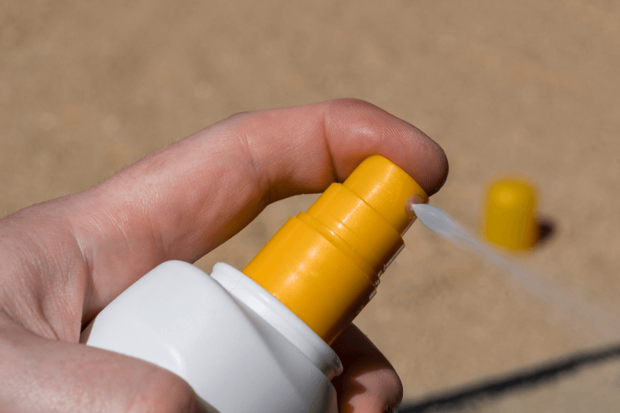This image shows a close-up of a hand using a spray bottle, likely sunscreen, with a yellow nozzle. A fine mist is being sprayed, and the blurred background suggests a sandy beach setting, reinforcing a sunny, outdoor context.