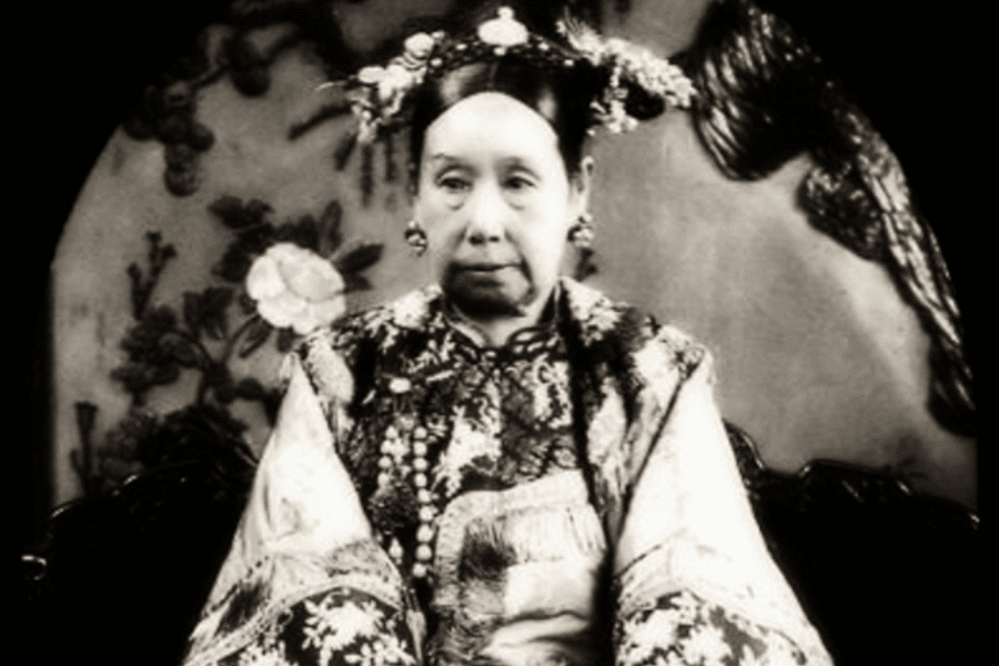 A black-and-white photograph of a woman in elaborate traditional Chinese attire, seated with a composed expression. She wears a richly embroidered robe adorned with intricate patterns and jewelry, and her hair is decorated with ornate headdresses featuring floral and beaded details. The background showcases a decorative screen with floral motifs, adding to the regal and dignified atmosphere.