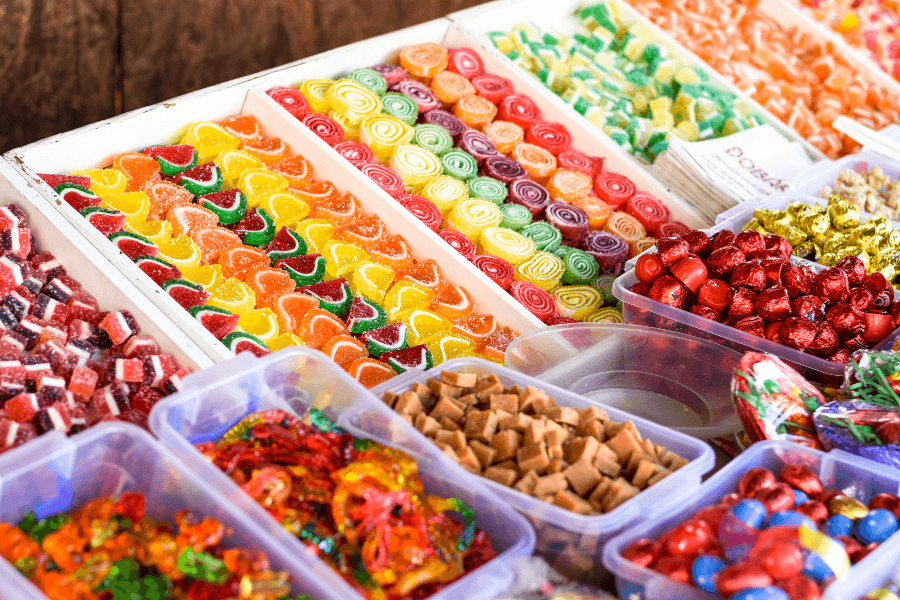 A market stall displaying a variety of colorful candies, including gummy fruit slices, spiral jelly candies, caramel cubes, and foil-wrapped chocolates. The sweets are arranged in neat rows and plastic containers, creating a vibrant and inviting display.