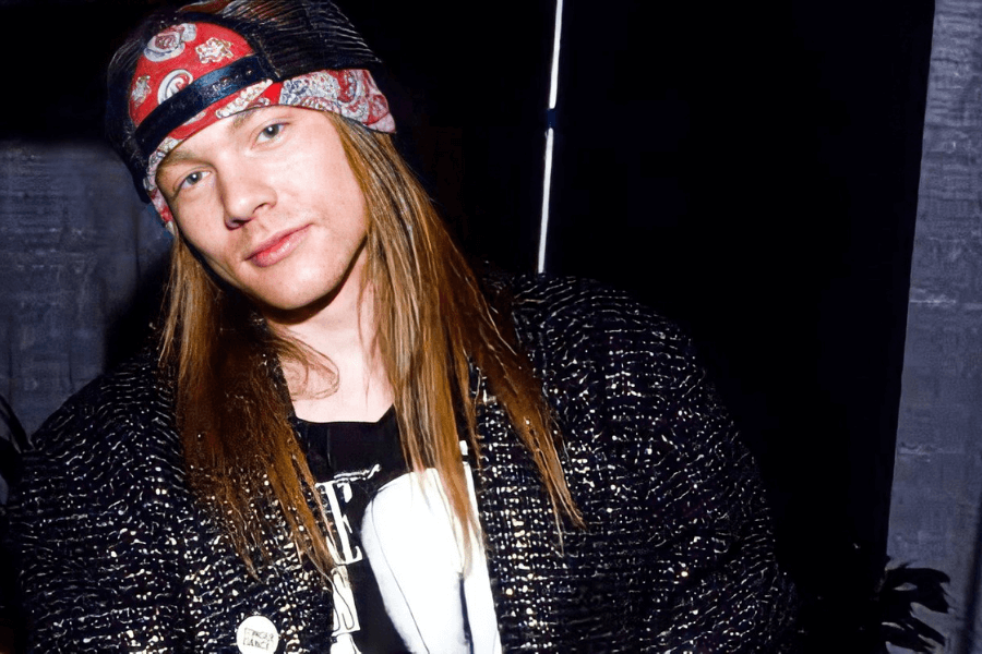 This image shows a man with long straight hair wearing a red patterned bandana, a textured black jacket, and a graphic t-shirt. He has a relaxed and casual expression, standing against a dark background, giving off a rock-and-roll or laid-back vibe.