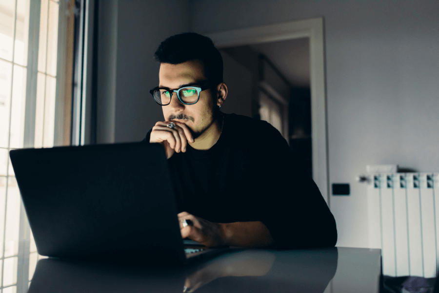 This image shows a man wearing glasses, sitting in a dimly lit room and focused on a laptop screen. His thoughtful expression, with his hand resting on his chin, suggests he is deeply engaged in work or study. The background includes a window and a radiator, enhancing the indoor setting.