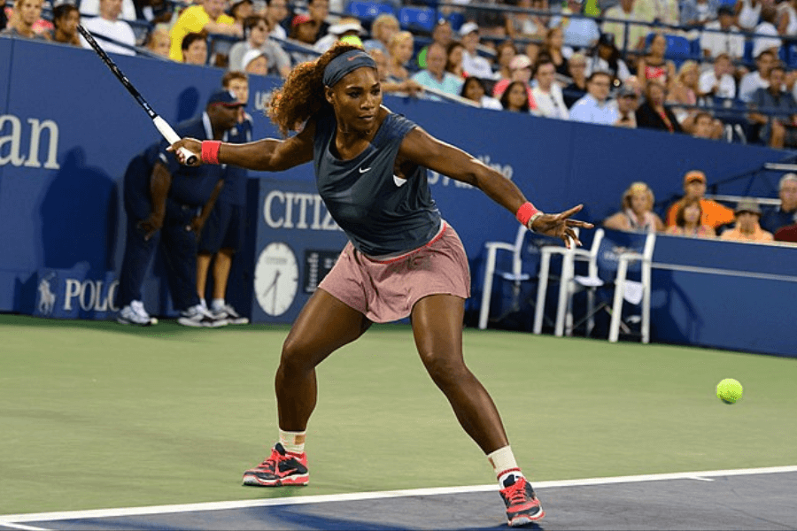 A female tennis player in athletic attire prepares to return a shot during a professional match. She is wearing a dark sleeveless top, a pink skirt, and matching pink and black sneakers, with her curly hair pulled back by a headband. The background shows a packed stadium with spectators watching the intense game.