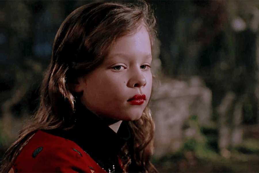 This image features a young girl with wavy brown hair and bright red lipstick, giving a serious or contemplative expression. She is wearing a red garment with black accents, set against a blurred, dark outdoor background that creates a mysterious or dramatic mood.