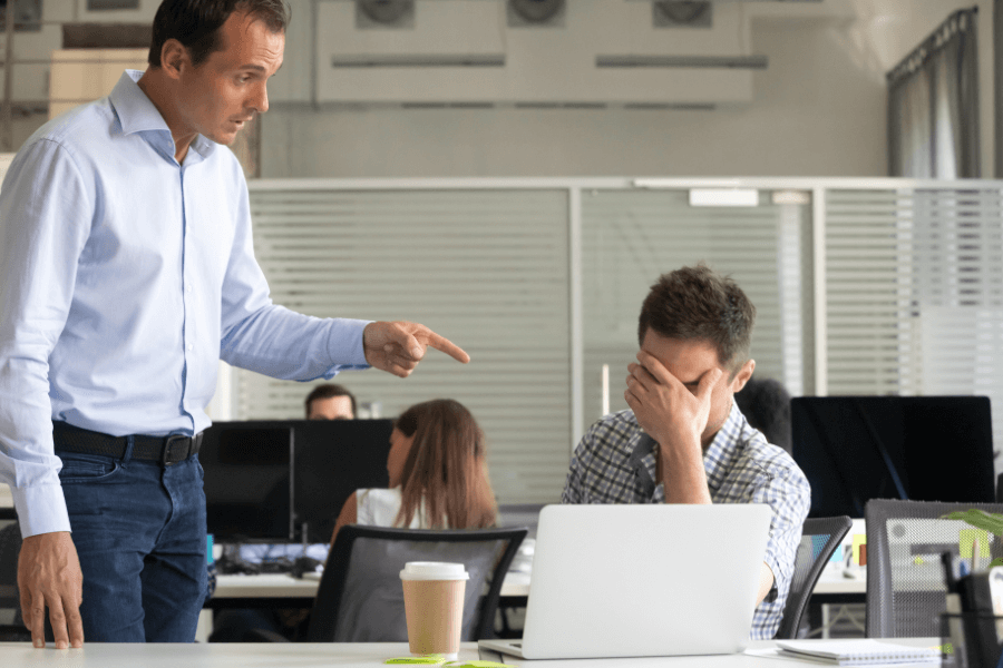 This image shows a workplace scenario where a man in a white shirt is pointing sternly at another man sitting at a desk, who has his hand over his face in frustration or distress. The background features an open office environment with desks, computers, and other employees working, adding to the professional setting.