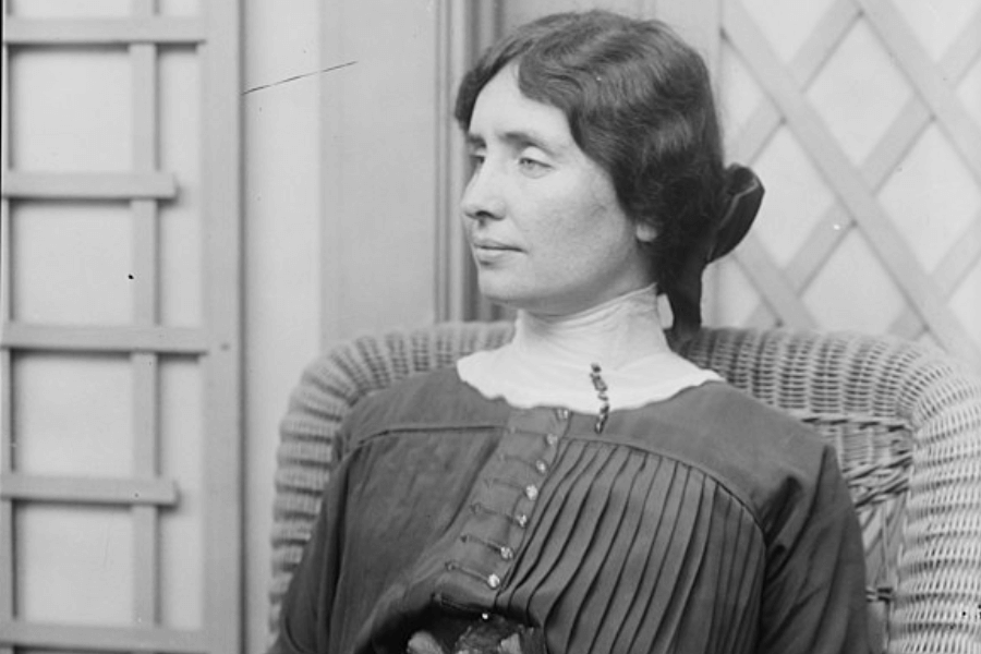 A black-and-white photograph of a woman seated in a wicker chair, gazing thoughtfully to the side. She wears a high-collared blouse with a pleated bodice, and her dark hair is pulled back with a ribbon. The background features a decorative lattice, adding to the serene and composed atmosphere.