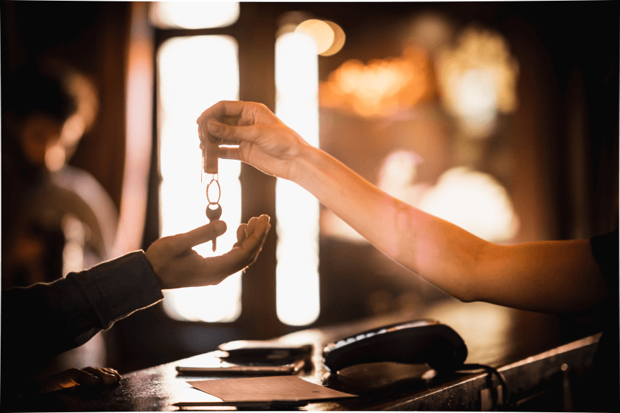 A hand extends a set of keys with a small keychain to another hand over a counter, with a payment terminal and clipboard visible nearby. The background is softly lit, suggesting a warm, indoor setting.