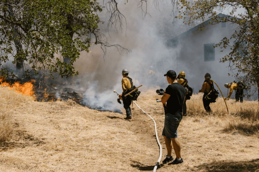 This image captures a scene of firefighters actively working to contain a brush fire near a house. Several firefighters in yellow protective gear are seen amidst smoke and flames in a dry, grassy area, while a person with a camera documents the event. The backdrop includes trees and a house, emphasizing the urgency of the situation.