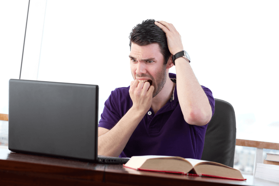 This image shows a man sitting at a desk with an open book and a laptop in front of him, displaying a distressed or anxious expression. He has one hand on his head and the other near his mouth, suggesting frustration or worry about something on the laptop screen. The bright setting and office environment enhance the focus on his emotional reaction.