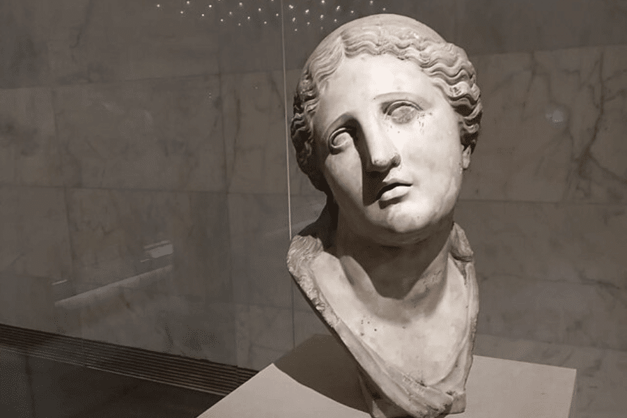 A marble bust of a woman displayed in a museum setting, with a serene yet sorrowful expression. Her head tilts slightly to one side, and her wavy hair is intricately carved, framing her face. The sculpture is set against a polished marble wall, with soft lighting casting shadows that highlight the delicate craftsmanship.