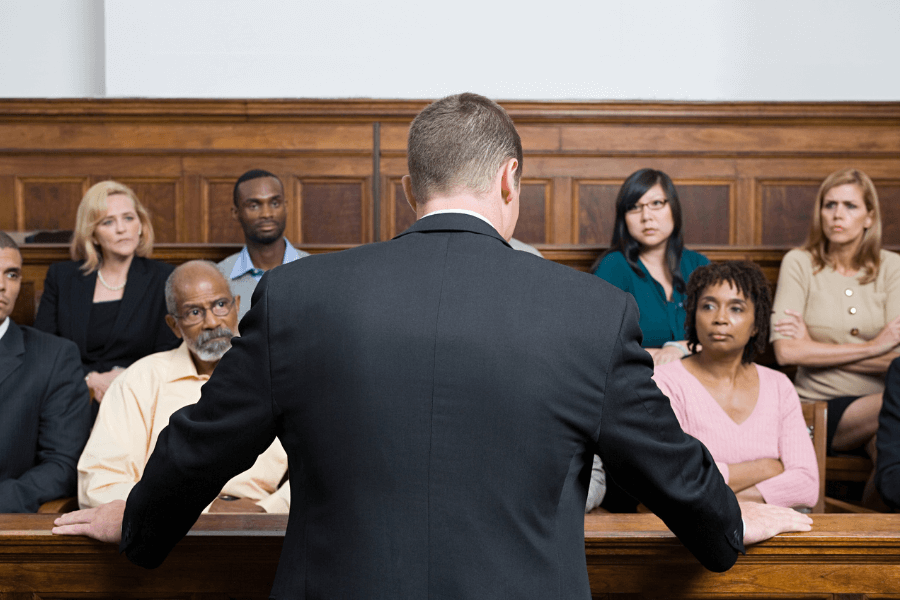 Man in a suit addressing a diverse group of jurors seated in a jury box, with serious and attentive expressions, inside a traditional wood-paneled courtroom.