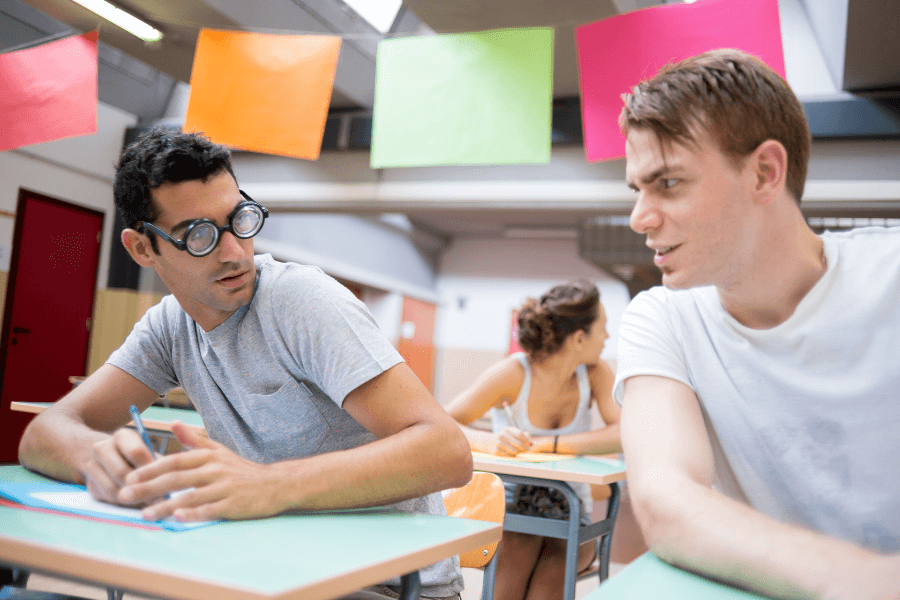 This image shows two young men sitting at desks in what appears to be a classroom. One man, wearing exaggerated round glasses, is holding a pen and looking suspiciously at the other, who seems to be speaking or interacting with him. The background includes another person working and colorful paper decorations hanging from the ceiling, creating a lively academic setting.