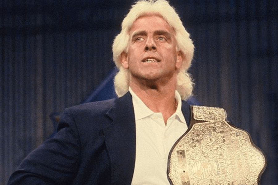 A man with blonde hair wearing a dark blazer and a white shirt poses confidently with a championship belt draped over his shoulder. The background is blurred, emphasizing his prominent stance.