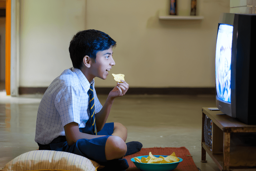 A young boy in a school uniform sits cross-legged on the floor, eating potato chips while watching television. The dimly lit room is illuminated by the TV screen, creating a cozy and focused atmosphere.
