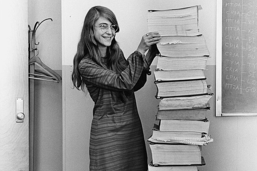A black-and-white photograph of a woman with long hair and glasses, smiling as she stands next to a towering stack of thick printed code books. She wears a striped dress and playfully holds the top of the stack, which appears to contain pages of computer programming. A chalkboard with technical notes is visible in the background.