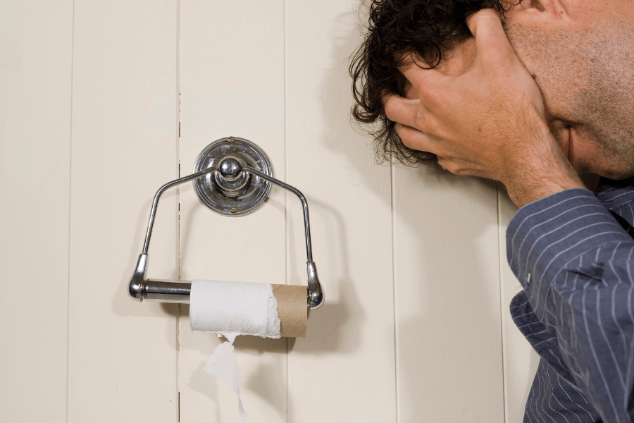 This image shows a man holding his head in apparent frustration or dismay next to an empty toilet paper holder with a cardboard roll still attached. The setting is a bathroom with light-colored paneling on the wall, and the situation humorously captures a relatable moment of inconvenience.