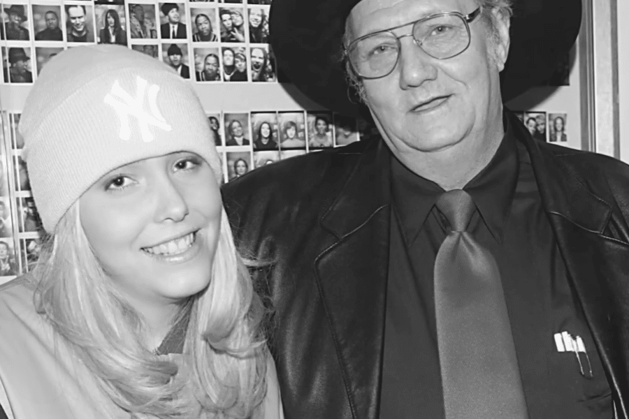 A black and white photograph of a young blonde woman wearing a New York Yankees beanie and a smiling expression, standing next to an older man in a black cowboy hat, glasses, and a leather jacket. The man wears a dark shirt and tie, with pens visible in his jacket pocket. Behind them, a wall covered in small photographs of people is visible.