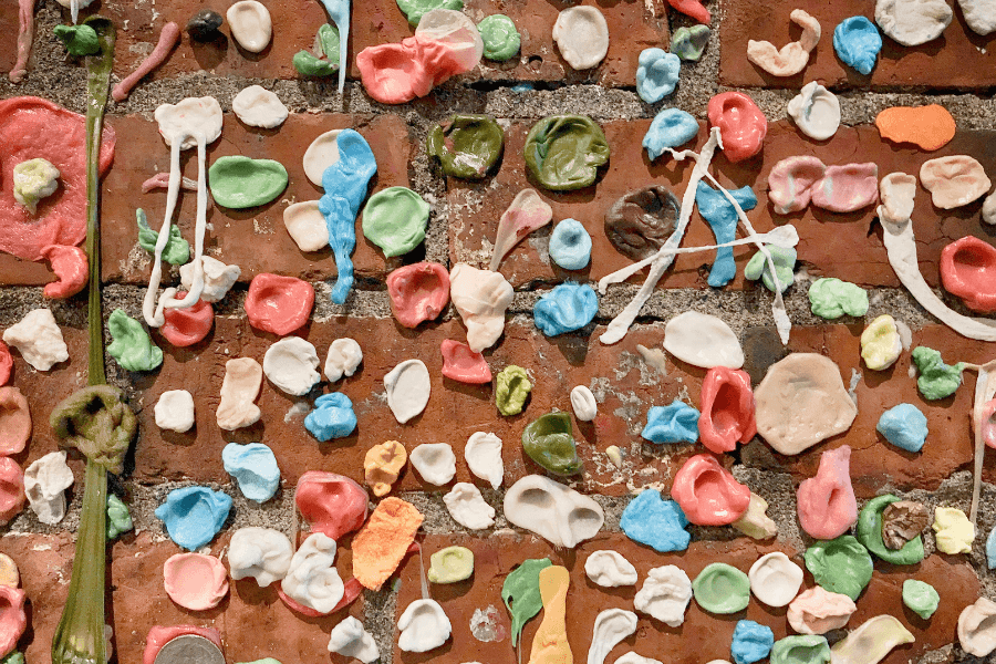 A close-up of a brick wall covered in colorful, chewed gum pieces in various shapes and sizes. Some gum pieces are stretched or layered, creating a textured and chaotic visual.