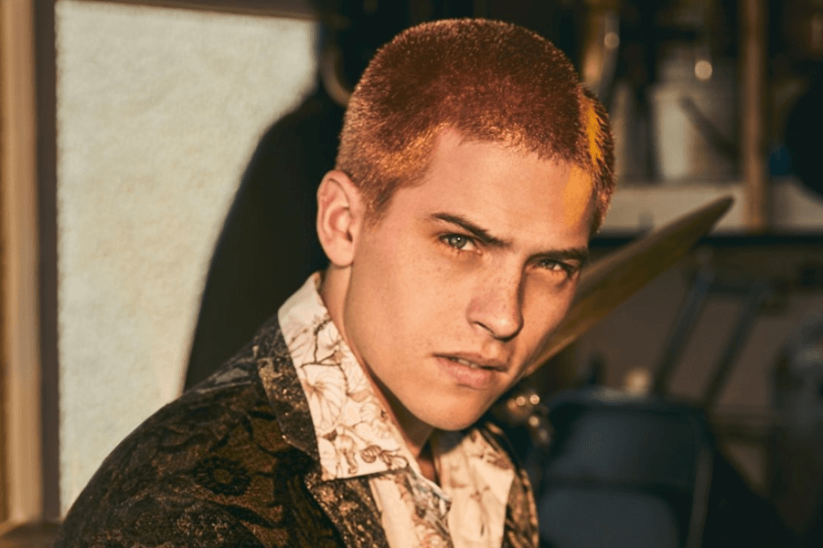 This image features a man with closely cropped, dyed hair and an intense gaze, wearing a patterned jacket over a floral shirt. The setting appears to be a dimly lit indoor space, with blurred objects in the background that suggest a workshop or creative environment. The lighting highlights his sharp features, giving the image a stylish and edgy feel.